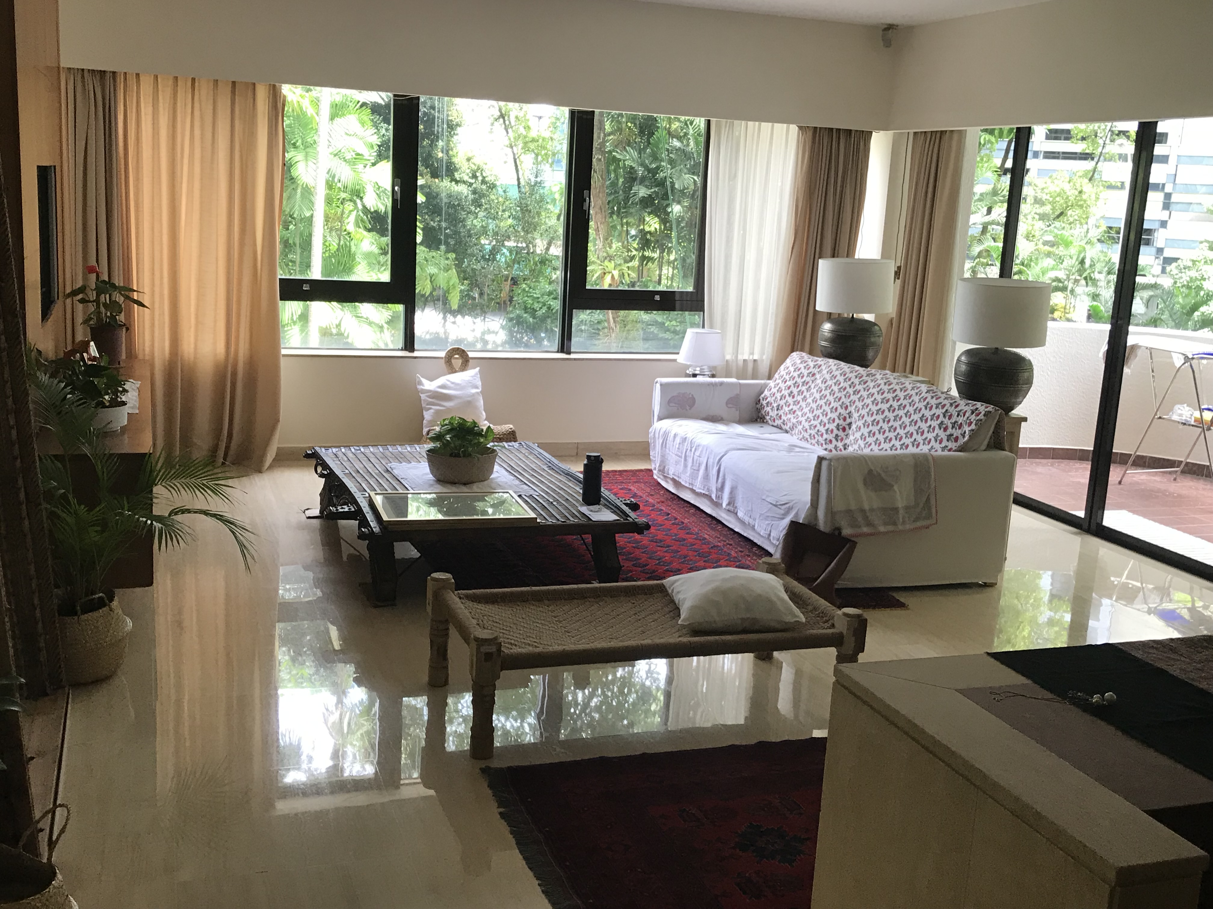 Singapore apartment — white sofa, charnpai charpoy, tropical canopy beyond the glass